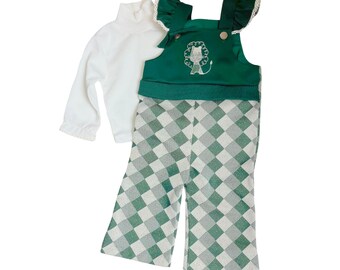 Op de afbeelding: Een witte coltrui met lange mouwen en een groen-witte geruite overall set. De overall top is groen met een leeuwenontwerp en ruches. De broek heeft een groen-wit geruit patroon.