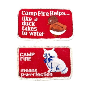 Op de afbeelding: Twee rode geborduurde patches met witte randen. De bovenste patch luidt "Camp Fire Helps... like a duck takes to water" met een eendenillustratie. De onderste patch luidt "CAMP FIRE means p-ur-rfection" met een kattenillustratie.