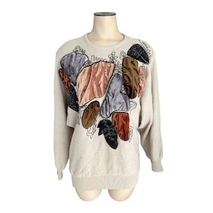 Può includere: Maglione beige con design applicato in velluto e ricamato colorato. Il maglione presenta toppe in velluto rosa, grigio, marrone e nero, con dettagli di ricamo nero. Il maglione ha un girocollo e maniche lunghe.