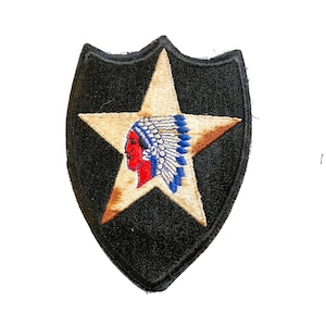 Puede incluir: Parche bordado negro con forma de escudo, con una estrella beige con un perfil nativo americano rojo y azul. El parche es probablemente un artículo militar o histórico. La estrella está delineada en marrón.