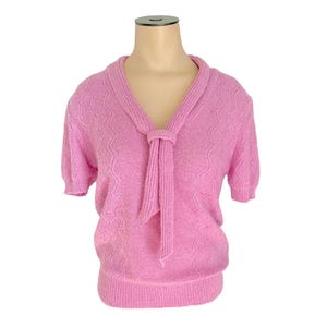 May include: A light pink short-sleeved sweater with a tie-neck design. The sweater has a textured pattern and a ribbed hem. The neckline is a V-shape, and the sleeves are short and slightly puffed.