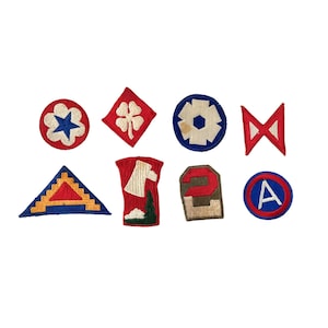 May include: A collection of eight vintage military patches in various shapes and colors. The patches feature designs such as a four-leaf clover, a star, a triangle, and the letter "A". Each patch is embroidered with detailed stitching.