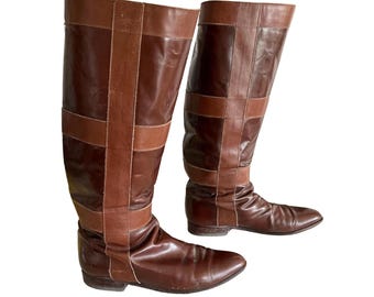 May include: A pair of knee-high brown leather boots with a pointed toe. The boots feature a darker brown leather accent design around the top and mid-calf. The boots are set against a white background.