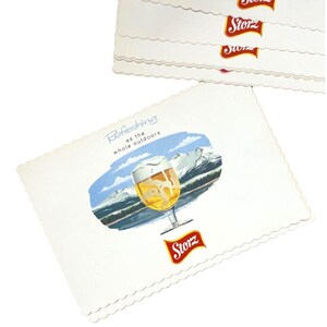 May include: A stack of rectangular white paper coasters with scalloped edges. Each coaster features a graphic of a beer glass against a mountain landscape, the text "Refreshing as the whole outdoors," and a red "Storz" logo at the bottom.