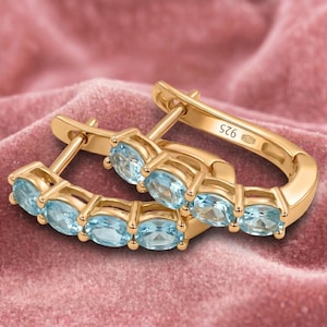 Könnte beinhalten: Ein Paar goldfarbene Creolen mit einer Reihe ovaler, hellblauer Edelsteine. Die Ohrringe sind vor einem rosa, samtigen Hintergrund platziert. Das Metall ist mit "925" gestempelt.