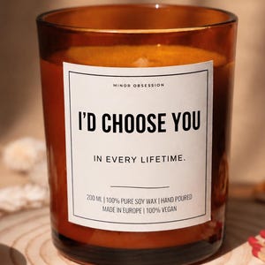 Può includere: Una candela da 200 ml in vetro ambrato con un'etichetta bianca riportante il testo Minor Obsession, I'd Choose You, In Every Lifetime, 100% Pure Soy Wax, Hand Poured, Made In Europe, 100% Vegan. La candela è riempita di cera arancione e uno stoppino.