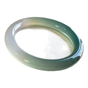 Puede incluir: Una pulsera tipo brazalete lisa y pulida hecha de piedra translúcida, que presenta una transición de color degradada desde un blanco lechoso pálido en un lado hasta un verde espuma de mar profundo y vibrante en el otro, con forma de banda continua.