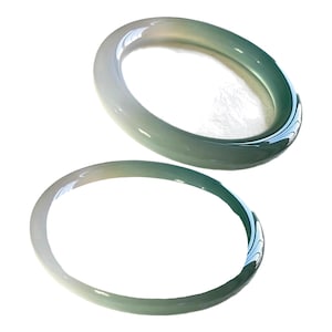 Puede incluir: Dos brazaletes rígidos lisos y pulidos hechos de piedra translúcida. Los brazaletes presentan una transición de color degradada, que cambia de un blanco lechoso pálido en un lado a un verde espuma de mar profundo y vibrante.
