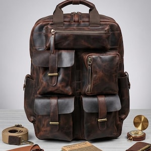May include: A dark brown leather backpack featuring a top handle, multiple exterior pockets with flap closures and brass hardware, and a zippered compartment. The backpack is surrounded by a compass, sunglasses, travel guides, and a notebook.