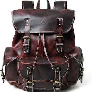 May include: A dark burgundy leather backpack featuring a top flap with a triangular logo, two front pockets, and two side pockets. The bag includes brass-toned metal buckles, white contrast stitching, and a drawstring closure under the main flap.