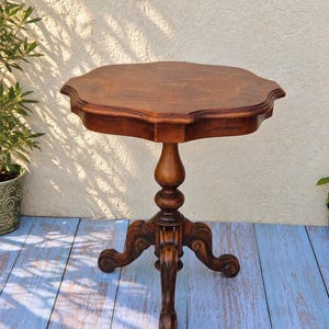 May include: A brown wooden pedestal table featuring a scalloped, multi-sided tabletop surface. The table is supported by a central turned column that transitions into a tripod base with three curved, ornate legs ending in scroll-shaped feet.