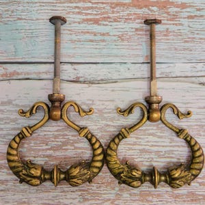 May include: Two antique brass drawer pulls with ornate, curved handles. The pulls have a detailed, textured design with a gold finish and a long threaded screw for mounting. The handles feature a decorative, stylized design.