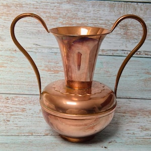 May include: A vintage copper pitcher with two curved handles. The pitcher has a bulbous base and a flared top. The copper has a warm, reddish-brown hue and shows signs of age. The handles are a darker shade of copper.