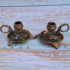 May include: A pair of matching copper-toned metal candle holders, each featuring a circular base with embossed scrollwork, a central candle cup, a curved side handle, and three small feet with a weathered, dark patina finish throughout.
