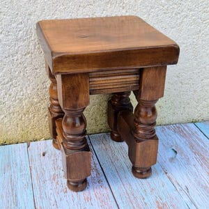 May include: A small, square wooden stool with a dark brown stained finish. The stool features a smooth, flat top surface and four legs with decorative turned detailing, including rounded bulbous sections and square-shaped feet at the base.