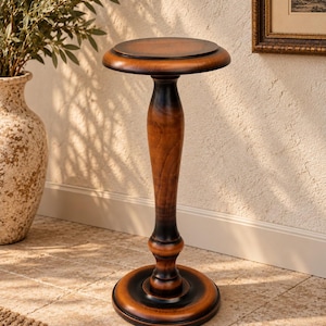 Peut inclure: Un piédestal haut et élancé en bois avec une finition brune polie et chaleureuse. Ce support présente un plateau circulaire, une colonne centrale tournée aux courbes lisses et une base circulaire large et stable posée au sol.