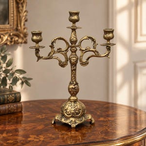 May include: A three-armed brass candelabra featuring an ornate, embossed floral and scrollwork design. The metal candle holder has a central stem with two symmetrical curved arms extending outward, each topped with a circular candle cup.