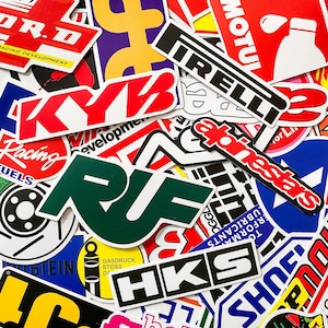 May include: A collection of colorful racing and automotive-themed stickers. The stickers feature various logos and text, including "KYB," "Pirelli," and "HKS." The stickers are in various shapes and sizes, with a mix of red, white, green, and black.