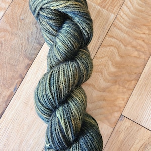 May include: A skein of yarn in a dark green and gold colorway. The yarn is made of wool and is perfect for knitting or crocheting.