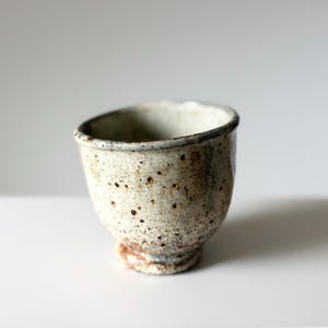 May include: A small, hand-thrown ceramic cup with a speckled off-white and beige glaze. The vessel features a rounded body, a slightly flared rim, and a distinct footed base with visible brown iron-rich spots across the textured surface.