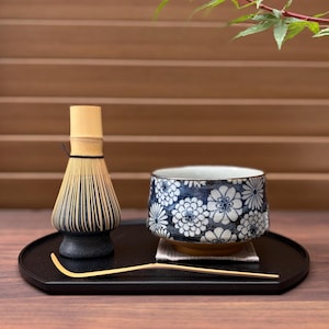 Può includere: Un frustino per matcha in bambù con base nera, una ciotola da tè in ceramica con motivo floreale blu e bianco e un sottile cucchiaino da tè in bambù. Questi oggetti sono disposti su un vassoio nero, con un piccolo panno quadrato sotto la ciotola.