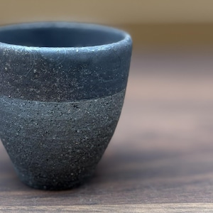 以下が含まれることがあります： 先細りの形状をした小さなダークグレーの陶器製カップ。上部は滑らかなマット仕上げで繊細な渦巻き模様があり、下部はざらざらとした粗い質感で、目に見える暗い粒子が表面全体に広がっています。