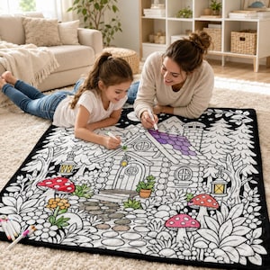 May include: A rectangular black and white fabric coloring blanket featuring a detailed line art illustration of a log cabin in a forest setting with mushrooms, trees, and lanterns. The blanket is partially colored with purple and green markers.