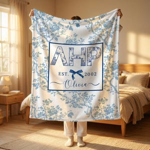 May include: A white fleece blanket featuring a blue floral pattern and a central rectangular frame. Inside the frame are the letters AHP, the text EST. 2002, a blue bow graphic, and the name Olivia in a cursive script font.