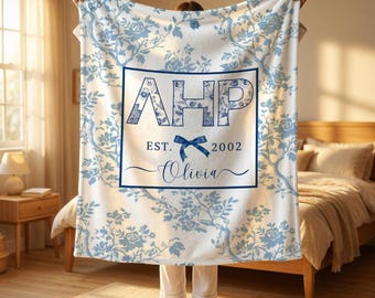 May include: A white fleece blanket featuring a blue floral pattern and a central rectangular frame. Inside the frame are the letters AHP, the text EST. 2002, a blue bow graphic, and the name Olivia in a cursive script font.
