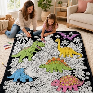 May include: A rectangular black and white dinosaur-themed coloring blanket featuring illustrations of a green T-Rex, a yellow long-necked dinosaur, a green Stegosaurus, a blue Triceratops, a pink Ankylosaurus, and a purple Pterodactyl.