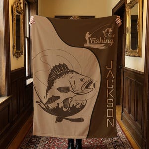 Puede incluir: Una manta marrón y beige con temática de pesca. El diseño presenta un pez saltando, una caña de pescar y la palabra "Fishing" con la silueta de un pescador. El nombre "Jackson" está impreso verticalmente.