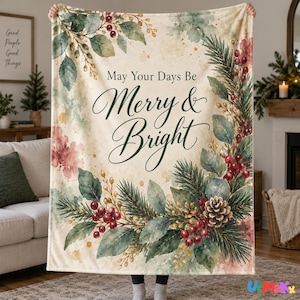 May include: A cream-colored throw blanket featuring a watercolor-style Christmas wreath design with pine branches, red berries, and a pinecone. The text "May Your Days Be Merry & Bright" is printed in the center in a dark green script font.