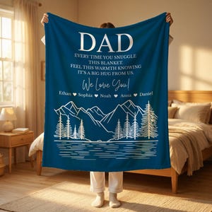 May include: A dark blue blanket with white text that reads "DAD" and "We Love You!" with names below. A mountain and lake scene is printed at the bottom. The blanket is being held up by a person.