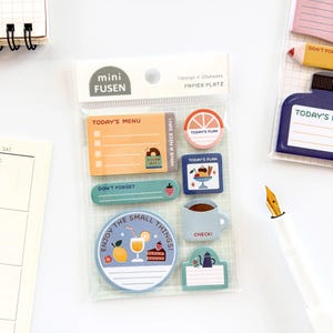 May include: A clear plastic package containing decorative sticky notes. The notes feature various designs, including a menu, a slice of orange, a coffee cup, and the text "Enjoy the small things!" and "Today's Plan".