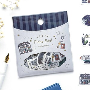 May include: A package of flake seals, featuring various illustrated stickers. The package has a blue and white striped top and a clear window showing the stickers. The stickers include house, key, and envelope designs. The text "Flake Seal" and "Papier Platz" are printed on the package.