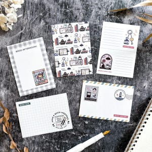 May include: A collection of decorative notepads with various designs and text. One notepad features a checkered pattern with "GOOD AFTERNOON!". Other notepads have illustrations of keys, clocks, and teapots, with phrases like "HAVE A NICE DAY!" and "GOOD MORNING!"