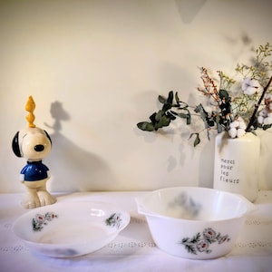 Op de afbeelding: Twee witte keramische kommen met bloemmotieven, naast een Snoopy-beeldje en een vaas met bloemen. De vaas heeft de tekst "merci pour les fleurs".
