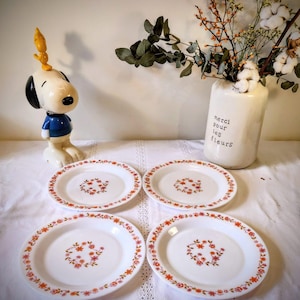 Puede incluir: Cuatro platos blancos con un diseño floral. Cada plato tiene un grupo central de flores rosas y un borde de flores naranjas y rosas. Los platos están sobre un mantel blanco. Una figura de Snoopy y un jarrón con flores también están en la imagen.