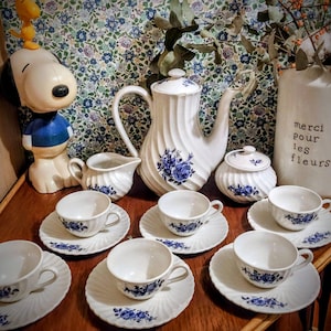 Könnte beinhalten: Ein weißes Porzellan-Teeservice mit blauen Blumenmustern, darunter eine Teekanne, eine Zuckerdose, ein Sahnekännchen und sechs Teetassen mit Untertassen. Eine Snoopy-Figur und eine Vase mit dem Text "merci pour les fleurs" sind ebenfalls vorhanden.