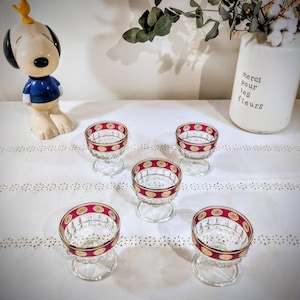 Peut inclure: Cinq coupes à dessert en verre transparent avec un bord rouge et doré à motifs sont disposées sur une nappe en dentelle blanche. Une figurine Snoopy et un vase blanc avec le texte "merci pour les fleurs" sont en arrière-plan.