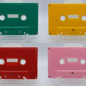 Può includere: Quattro musicassette colorate, verde, giallo, rosso e rosa. Ogni cassetta ha una forma rettangolare con angoli arrotondati, due fori circolari e un'apertura rettangolare centrale. Le cassette sono esposte su supporti trasparenti su sfondo bianco.