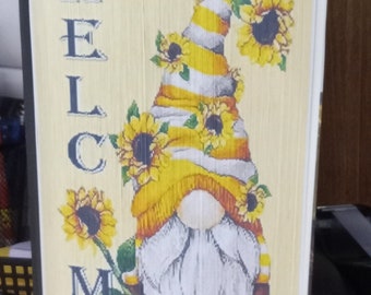 May include: A decorative book featuring a gnome design. The gnome has a yellow and white striped hat decorated with sunflowers. The word "WELCOME" is printed in navy blue on the book's cover. The gnome holds a sunflower.