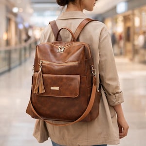 Puede incluir: Mochila de cuero sintético marrón con múltiples compartimentos, cremalleras y un bolsillo frontal. La bolsa tiene un asa superior, correas ajustables y una borla decorativa. Los herrajes son dorados.
