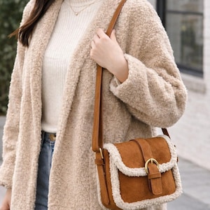 Puede incluir: Un abrigo de peluche color canela y un bolso bandolera de ante marrón con hebilla dorada. El bolso tiene un ribete color crema. La persona lleva un jersey color crema y vaqueros azules.