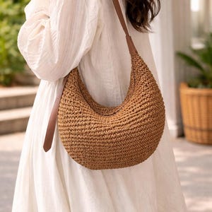 May include: A crescent-shaped shoulder bag in a woven brown material, featuring a brown leather strap. The bag has a textured surface and is worn over the shoulder. The neutral colour of the bag complements the white dress.
