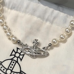 May include: A pearl necklace with a silver pendant featuring a planet design encrusted with clear crystals. The necklace is displayed on a white fabric surface.