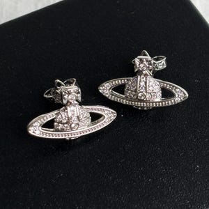 Puede incluir: Un par de pendientes de botón plateados. Cada pendiente presenta un diseño similar a un planeta con un anillo y una corona, incrustado con pequeños cristales brillantes. Los pendientes se muestran sobre una superficie negra.