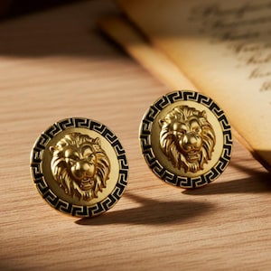 May include: Two gold-coloured cufflinks featuring a lion's head motif. Each cufflink has a black border with a Greek key design. The background includes a vintage paper and a leather-bound book.