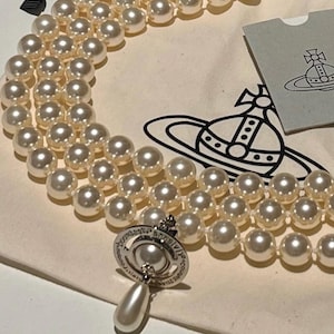 May include: A cream-colored pearl necklace with three strands, featuring a silver clasp and a pendant with a pearl drop. The necklace is displayed on a cream-colored pouch with a black ribbon and a tag with a logo.