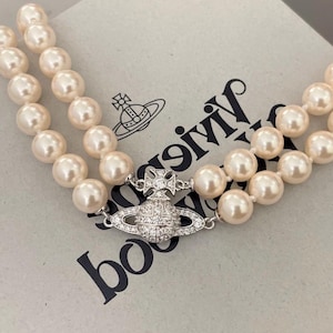 Puede incluir: Un collar de perlas de doble hebra con un cierre plateado. El cierre presenta un diseño de orbe decorativo con una cruz, incrustado con pequeños cristales transparentes. Las perlas son de color crema.